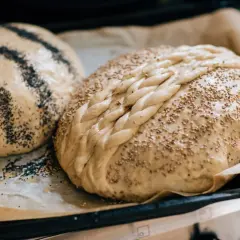 Två brödlimpor på bakplåtspapper. Den ena är flätad och toppad med sesamfrön, den andra har vallmofrön i ränder. Surdeg bubblar inte, men dessa ser redo ut för ugnen.