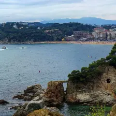Widok na zatokę z łodziami i plażą. W oddali widać miasto i góry. Pogoda na Costa Brava zapowiada się na spokojną.