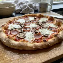 En gyllene, hemmagjord pizza med skinka och champinjoner ligger på en träskärbräda.