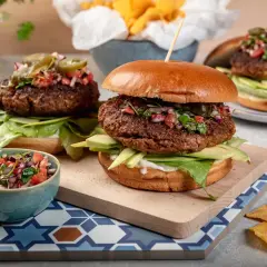 Dwa soczyste burgery wołowe z awokado, salsą i jalapeno, obok miseczki z salsą i nachos.