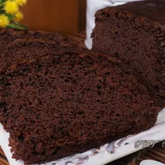 Plumcake al cacao sofficissimo, affettato su un piatto bianco, con fiori gialli sullo sfondo.