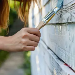 Malowanie drewnianej elewacji. Pędzel z jasnym kolorem nakłada farbę na deski. Dowiedz się, czym pomalować olejowane drewno, by odnowić jego wygląd.