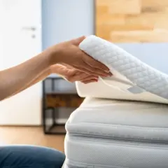 Ręce układają biały, pikowany topper na łóżko, poprawiając komfort snu.