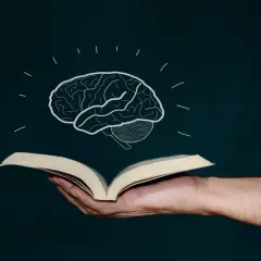 Mano sostiene libro abierto, de él surge un cerebro dibujado, símbolo de la lectura global y el conocimiento.
