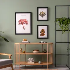 Salon z zielonymi, ciepłe kolory ścian do salonu. Obok fotel, drewniana komoda z dekoracjami i grafiki z motywami morskimi.
