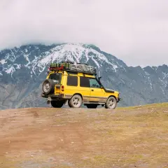 Żółty off road 4x4 z bagażem na dachu i zapasowym kołem na tyle, zaparkowany na zboczu góry, pod ośnieżonymi szczytami.