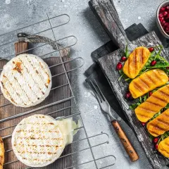 Dwa krążki serów na grilla, jeden lekko roztopiony, obok grzanki. Na desce obok grillowane kawałki dyni z rukolą i granatem.