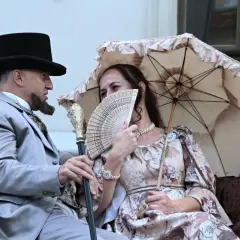 Para w strojach z epoki, mężczyzna w cylindrze i z laską, kobieta z wachlarzem i parasolką.