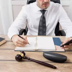 Karnista kto to? Prawnik w białej koszuli i krawacie siedzi przy biurku, pisze w notatniku, trzymając telefon. Obok leży młotek sędziowski i waga.
