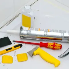 Werkzeuge zum Silikon von Fliesen entfernen: Kartuschenpresse, Cutter, Schaber, Bürste und eine Flasche mit Reiniger.