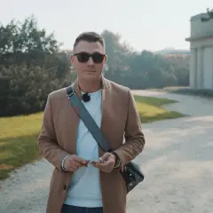 Mężczyzna w beżowym płaszczu i okularach przeciwsłonecznych, trzymający klucze, przygotowuje się do kursu fotografii telefonem w parku.