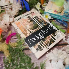 Książka "Pociąg Nr 13" Renaty Wasilewskiej, ozdobiona perłami i piórami, niczym oryginał oprócz błękitnego nieba, otoczona kwiatami.