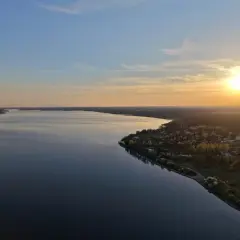 Zachód słońca nad Zalewem Sulejowskim. Spokojna tafla wody odbija niebo, a na brzegu widać domki letniskowe i las.
