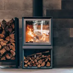 Płonąca koza na tle kamiennej ściany. Obok stojak z drewnem, a pod piecem schowek na opał.