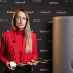 Kobieta w czerwonej marynarce prezentuje element grzałki elektrycznej, kluczowy dla sprawności grzałki elektrycznej. Obok stoi bojler.