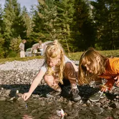 Två barn leker med pappersbåtar vid en sjö. I bakgrunden syns ett tält och träd. En härlig 5 kamp fest i naturen.