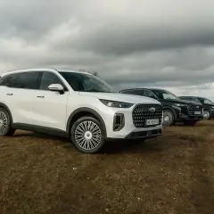 Biały Jaecoo 9 i dwa ciemne SUV-y na wzgórzu pod pochmurnym niebem.