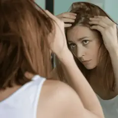 Mujer preocupada revisa su cuero cabelludo en el espejo, buscando soluciones sobre como frenar la caida del cabello.