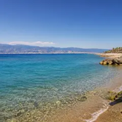 Spiaggia di Reggio Calabria con mare cristallino, palme e case bianche. All'orizzonte, montagne azzurre sotto un cielo limpido.