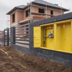 Żółta skrzynka z instalacją gazową, widoczna na tle budowanego domu i ogrodzenia.