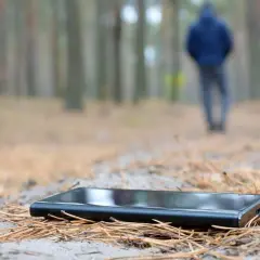 Ein Handy verloren, was tun? Ein schwarzes Smartphone liegt im Wald auf Nadeln. Im Hintergrund geht eine Person weg.