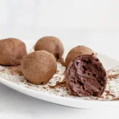 Cinco trufas de chocolate espolvoreadas con cacao en polvo sobre un plato blanco. Una trufa está partida, revelando su interior cremoso.
