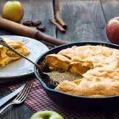 Una deliziosa torta di mele in padella, appena sfornata, con una fetta servita su un piatto. Mele fresche e cannella completano la scena rustica.