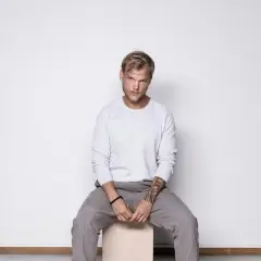 Avicii, siedzący na stołku, w szarym swetrze i spodniach. Jego śmierć wciąż budzi pytania.