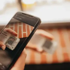 Ręce trzymają smartfon, który skanuje kod QR na małym urządzeniu. To może być część procesu śledzenia przesyłki przez kuriera Hellmann.