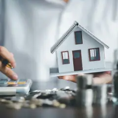 Osoba liczy koszty, trzymając model domu. W tle słoik z monetami i stosy pieniędzy, co sugeruje plan gospodarczy wspólnoty mieszkaniowej.