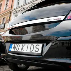 Czarne auto z indywidualnymi tablicami rejestracyjnymi "NO KIDS" na tle kamienicy.