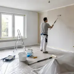 Malowanie pokoju: mężczyzna wałkiem odświeża ściany. Na podłodze folia ochronna, drabina i wiaderka z farbą.