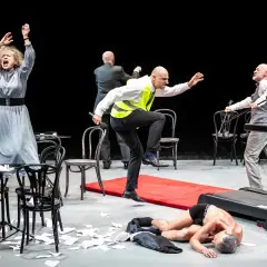 Scena teatralna z kobietą w szarej sukni, mężczyzną w kamizelce odblaskowej i leżącym na bieżni. Czy to symbol tego, ile trwają przerwy w teatrze?