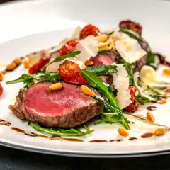 Saftiges Tagliata di Manzo mit Rucola, Kirschtomaten, Pinienkernen und Parmesan auf einem weißen Teller.