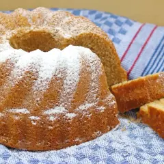 Puszysta babka panettone, oprószona cukrem pudrem, leży na niebiesko-białym ręczniku. Obok niej leżą dwa kawałki ciasta.