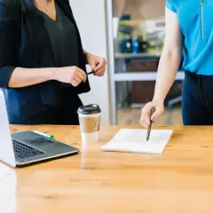 Zwei Frauen besprechen Dokumente am Tisch. Ein Laptop und Kaffee stehen bereit. Möglicherweise geht es um die Auszahlung Lebensversicherung Steuer.