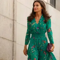 La reina Letizia, con un vestido verde estampado y pendientes llamativos, camina con elegancia junto a un muro de piedra.