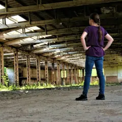 Dziewczynka w jeansach i fioletowej bluzce stoi w opuszczonej hali, odkrywając tajemnice urbex.