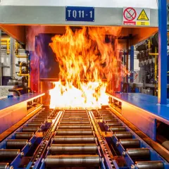 Płomienie buchają z pieca, w którym odbywa się nawęglanie stali. Taśmociąg z rolkami transportuje metal przez gorący proces.