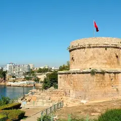 Antalya Altstadt: Ein runder Steinturm mit türkischer Flagge überragt die Bucht. Im Hintergrund moderne Gebäude und Felsen.