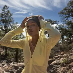 Une femme ajuste son bandeau et sa veste légère, parfaite pour savoir comment s'habiller pour une randonnée en été. Paysage rocheux et arbres en arrière-plan.