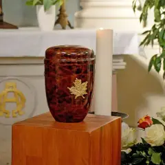Elegancka urna z prochem człowieka, ozdobiona złotym liściem klonu, stoi obok białej świecy i bukietu róż.