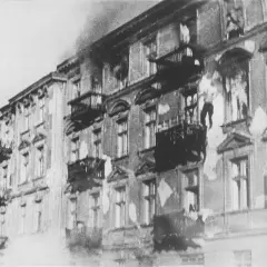 Brennende Gebäude im Warschauer Ghetto. Rauch steigt auf, Fenster sind zerbrochen, Balkone sind dunkel.