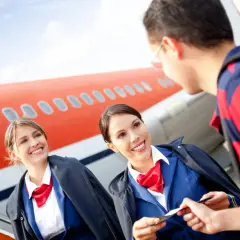 Zwei lächelnde Flugbegleiterinnen im blauen Anzug mit roten Halstüchern prüfen eine Bordkarte vor einem Flugzeug. Die Ausbildung zur Flugbegleiterin und das Gehalt sind attraktiv.