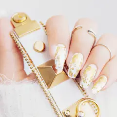 En hand med vita naglar dekorerade med guld nagellack, som en lyxig touch. En vit och guldig accessoar syns också.