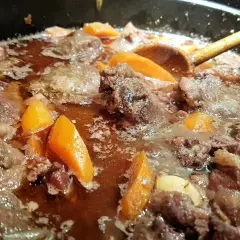 Un plat mijoté de joue de porc avec des carottes, dans une sauce brune savoureuse. Une cuillère en bois remue doucement.
