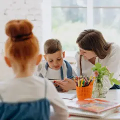 Nauczycielka pokazuje chłopcu obrazek w książce, ucząc go angielskiego. Obok stoi dziewczynka.