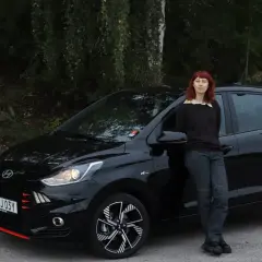 En kvinna med rött hår lutar sig mot en svart Hyundai i10. Bilen är redo för ett test.