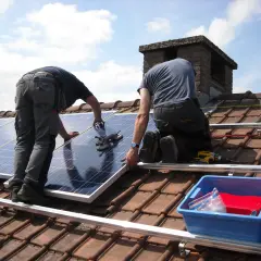 Dwóch mężczyzn montuje panele PV na dachu. Widać narzędzia i niebieskie pudełko z akcesoriami.