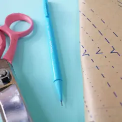 Pink scissors and a blue pen lie next to a piece of pattern paper, ready for a project to place on fold.